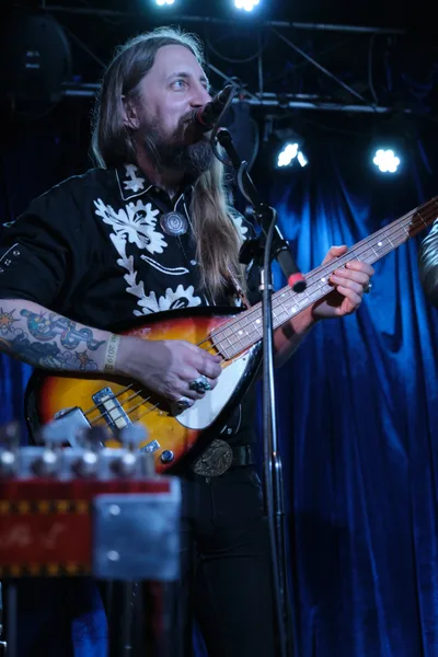 Tobias Berblinger of Roselit Bone delivers an intense performance at Dante's in Portland, his weathered hands commanding a sunburst bass while crystalline stage lights pierce the intimate venue's darkness. The long-haired musician's black shirt adorned with white floral patterns creates striking contrast against the deep blue backdrop, while intricate tattoos snake down his forearms. His upward gaze and focused expression capture a moment of musical transcendence during RX Fest 2019.