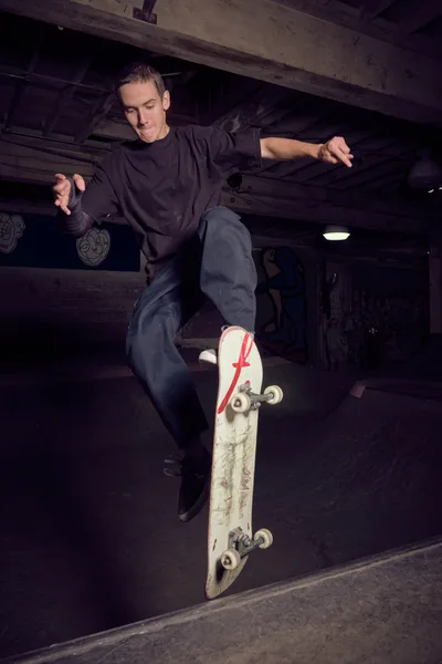 A young skateboarder executes a fluid carve through the concrete bowl at Commonwealth Skateboarding in Portland's Buckman neighborhood. The moody lighting catches the weathered skateboard bearing a distinctive red 'F' logo as the rider maintains perfect balance through the curved transition. Exposed wooden beams overhead and the industrial atmosphere create an authentic underground skate culture scene, with the worn concrete and ambient shadows emphasizing the raw energy of street skating.