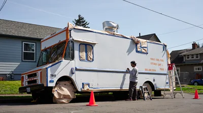 A muralist works diligently on Papa's Soul Food Kitchen Cart in Portland's King neighborhood, surrounded by residential houses and spring greenery. The artist, wearing a white long-sleeved shirt and baseball cap, stands beside the white food truck adorned with orange and blue trim, methodically applying paint while orange traffic cones mark the workspace. Afternoon light casts gentle shadows across the suburban street scene, capturing the quiet transformation of a neighborhood business through public art.