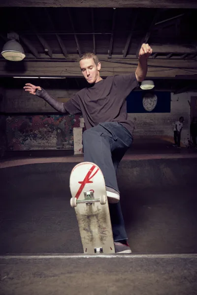 A focused skateboarder sits contemplatively on weathered concrete steps in the shadowy depths of Commonwealth Skateboarding in Portland's Buckman neighborhood. Arms outstretched in perfect balance, he embodies the zen-like concentration that defines bowl skating culture. The stark industrial lighting cuts through the underground atmosphere, illuminating weathered walls adorned with graffiti while his skateboard—marked with a distinctive red 'F' logo—rests against the worn concrete, telling stories of countless sessions in this subterranean sanctuary.
