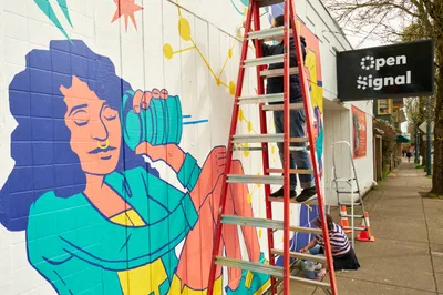 Two muralists work on a vibrant public art piece titled "Let's Talk" on the exterior wall of Open Signal community media center on Martin Luther King Jr. Boulevard in Portland's Eliot neighborhood. One artist perches on a red stepladder while painting detailed sections of the colorful mural, which features a figure with flowing blue hair holding turquoise objects against a backdrop of geometric patterns in coral, yellow, and teal. The scene captures the collaborative spirit of community art-making under overcast Pacific Northwest skies.