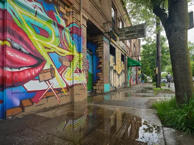 Vibrant street art transforms the brick facade of the former Oregon Theater on Southeast Division Street, where bold graffiti featuring crimson lips and electric yellow lettering creates a striking contrast against weathered brown brick. Puddles on the cracked sidewalk mirror the colorful murals while a solitary pedestrian passes beneath the canopy of mature street trees. The scene captures Portland's Richmond neighborhood where urban decay meets artistic expression, with the vintage tavern sign and rain-slicked pavement telling stories of the district's evolving character.