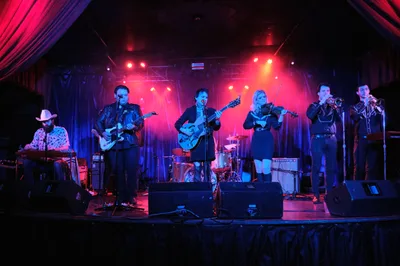 Six musicians from Roselit Bone command the intimate stage at Dante's in Portland, bathed in saturated crimson and azure theatrical lighting. The ensemble features electric guitars, violin, brass instruments, and keyboards, creating a dynamic country-fusion performance. Dramatic spotlights pierce through the smoky venue atmosphere, casting the performers in theatrical silhouettes against draped curtains and exposed stage rigging.
