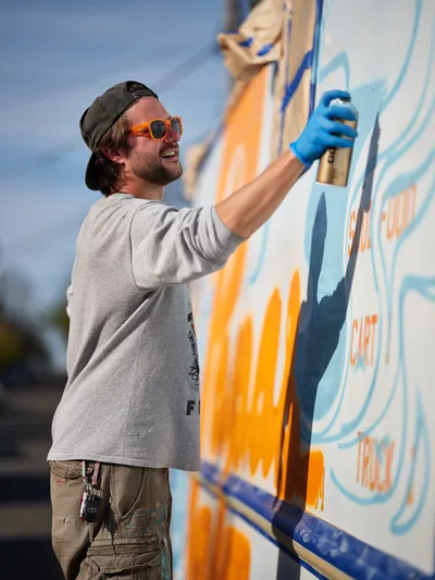 A bearded muralist in orange sunglasses and backwards cap applies bright blue paint to a colorful food truck exterior in Portland's King neighborhood. The artist works with focused concentration, his paint-stained cargo pants and blue latex gloves testament to hours of creative labor. Orange and blue hues dominate the emerging artwork against the soft afternoon sky, while the food truck's white canvas transforms under his skilled brushstrokes.