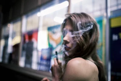 A young woman with flowing brown hair exhales vapor while positioned against the blurred backdrop of a convenience store on Northeast 7th Avenue in Portland's Irvington neighborhood. The shallow depth of field creates an intimate portrait where colorful storefront signage dissolves into soft bokeh, emphasizing her contemplative profile. Natural daylight illuminates her bare shoulder and the translucent vapor cloud, capturing a moment of urban solitude amid the commercial streetscape.