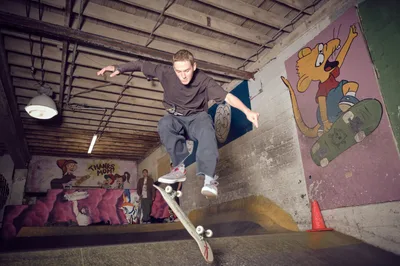 A skateboarder executes a dynamic aerial maneuver in the concrete bowl at Commonwealth Skateboarding in Portland's Buckman neighborhood. The exposed wooden beam ceiling and weathered industrial pendant lights frame the action, while vibrant cartoon murals featuring animated characters create a nostalgic backdrop. The rider, caught mid-flight in baggy street wear, embodies the raw energy of underground skate culture against the gritty urban canvas of this Southeast Portland institution.