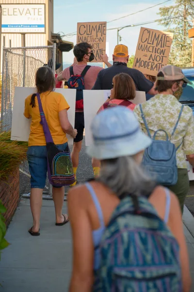 A determined group of climate activists gathers on the sidewalk outside Representative Kurt Schrader's office in Oregon City, their handwritten cardboard signs declaring "Climate Inaction Is Not Moderate." Golden afternoon light filters through the suburban setting as protesters of various ages stand unified along McLoughlin Promenade, some wearing masks and carrying colorful bags, creating an intimate yet powerful scene of grassroots political engagement. The composition captures both the urgency of their message and the everyday nature of democratic participation in small-town America.