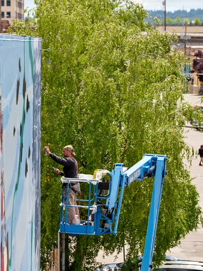 Two artists, Jeremy Nichols and Devin Finley, work from a blue aerial lift platform to create a large-scale mural on the side of a building in Southeast Portland. The scene captures them mid-creation against a backdrop of spring foliage from a mature tree, with urban brick architecture visible in the frame. Bright midday sunlight illuminates their concentrated work on the Portland Street Art Alliance project, highlighting the intersection of public art and community transformation in Portland's Central Eastside district.