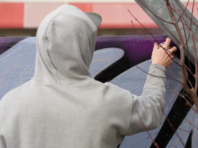 Travis Fields, known as Campographic, works intently on a vibrant mural at the Clay Creative Building in Portland's Hosford-Abernethy neighborhood. Wearing a gray hoodie, the artist reaches toward his canvas of purple, blue, and black abstract forms painted on weathered concrete. The intimate perspective captures the focused concentration of street art creation, with dried branches adding organic texture to the urban creative process.