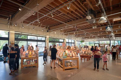 Inside Powell's City of Books in Portland's Pearl District, warm afternoon light streams through expansive windows, illuminating browsing customers amid carefully arranged wooden display tables. The industrial-chic ceiling reveals exposed beams and track lighting above the bustling literary sanctuary, where families and book lovers of all ages navigate between organized displays of featured titles. The polished concrete floors reflect the gentle glow of the space, creating an inviting atmosphere that captures the essence of Portland's renowned independent bookstore culture.