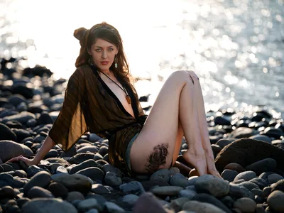A woman with dark brown hair sits contemplatively among weathered river rocks along Oregon's Columbia River Gorge, her olive-toned wrap flowing around her figure. The late afternoon sun creates dramatic backlighting, casting her silhouette against sparkling water and transforming the rocky riverbed into a canvas of warm shadows and brilliant highlights. Her pendant necklace and dangling earrings catch the ethereal light as she gazes toward the camera with a serene, introspective expression.