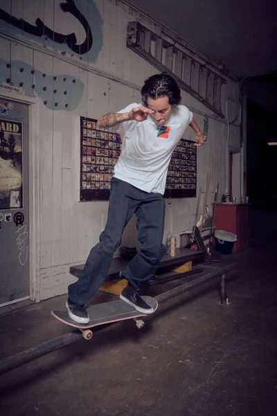 A tattooed skateboarder in a white tee and dark jeans executes a smooth ride through the weathered interior of Commonwealth Skateboarding in Portland's Buckman district. The moody lighting illuminates vintage skate memorabilia plastered across white wooden walls, while the rider's focused expression and fluid motion capture the authentic spirit of skate shop culture. Shadows dance across the worn floorboards as the session unfolds in this iconic Southeast Portland skate sanctuary.