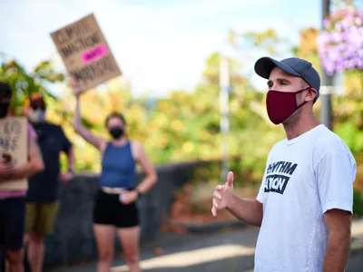 Organizer Peter Marks, wearing a burgundy face mask and gray baseball cap, gestures emphatically while speaking to assembled protesters outside Representative Kurt Schrader's office in Oregon City. The afternoon light casts warm tones across the scene as blurred demonstrators hold signs in the background, creating an atmosphere of peaceful civic engagement. His white t-shirt bearing partial text suggests grassroots activism, while the suburban setting frames this moment of political discourse.