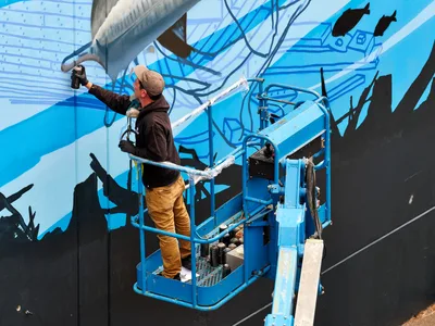 Jeremy Nichols and Devin Finley work from a blue scissor lift to paint an expansive mural featuring flowing blue forms and black silhouettes on a building wall in Southeast Portland's Central Eastside. The artist in mustard-colored pants extends his spray can toward the dynamic composition while bottles of paint rest in the lift's platform. Crisp daylight illuminates the industrial setting where street art transforms urban architecture.