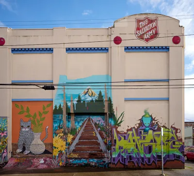 Vibrant street art adorns the weathered beige facade of a former Salvation Army warehouse in Portland's Central Eastside district. The commissioned murals create a striking narrative across the building's lower level, featuring a whimsical gray cat among botanical elements, a wooden pier stretching toward distant mountains, and bold graffiti-style lettering in electric green and purple. Power lines slice horizontally through the frame against a backdrop of azure sky and drifting clouds, emphasizing the gritty urban transformation of this industrial neighborhood.