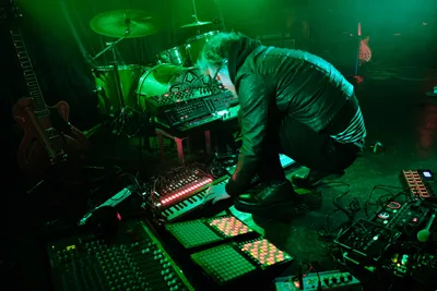 Electronic musician Eli Savage, performing as ellipsism, hunches intently over a sprawling array of synthesizers, drum machines, and MIDI controllers during his live set at RXFest 2019. Bathed in otherworldly emerald stage lighting, the artist manipulates his electronic instruments with focused precision while surrounded by the intimate darkness of Dante's venue in Portland's downtown core. The atmospheric green wash transforms the performance space into a cyberpunk dreamscape, highlighting the glowing control surfaces and creating dramatic shadows across the cluttered stage floor.