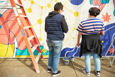 Two muralists work on the vibrant "Let's Talk" public art installation at Open Signal in Portland's Eliot neighborhood. One artist in a navy vest and hoodie stands contemplatively before the wall while another in a striped shirt and headphones actively paints the colorful geometric design. The mural features bold yellow molecular patterns, coral and teal geometric shapes, and dynamic blue elements against a white concrete wall, with an orange stepladder and paint supplies scattered across the concrete sidewalk.