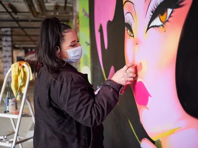 Multidisciplinary artist Vilatose applies finishing touches to a luminous portrait mural in the industrial confines of Clay Creative's parking garage in Portland's Hosford-Abernethy neighborhood. Wearing a protective mask and dark work attire, she delicately airbrush details onto the large-scale feminine face rendered in electric pinks, corals, and sunset hues. The contrast between the raw concrete surroundings and the radiant artwork creates a compelling dialogue between urban grit and artistic transformation.