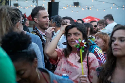 A jubilant crowd gathers at White Owl Social Club during Your Sunday Best's season opener, their faces illuminated by warm string lights overhead. Festival-goers sport vibrant tie-dyed shirts and casual summer attire, creating an intimate tapestry of celebration in Portland's beloved outdoor venue. The shallow depth of field captures genuine moments of connection as friends laugh and socialize in the golden hour glow.