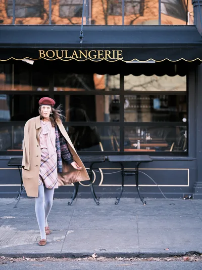 A young woman in a burgundy beret and camel coat strikes a dynamic pose outside a boulangerie on Portland's Alberta Street. Her wind-swept hair and layered ensemble of plaid scarf, patterned sweater, and white tights create an effortlessly Parisian aesthetic against the urban backdrop. The golden afternoon light catches the brass lettering of the French bakery behind her, while outdoor café chairs hint at European-style street life in this Northeast Portland neighborhood.