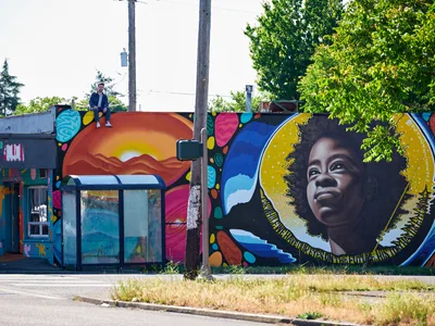 Travis Fields sits triumphantly atop a weathered building in Portland's Alberta district, having completed a vibrant mural celebrating poet Amanda Gorman. The large-scale artwork features Gorman's portrait rendered with warm golden tones against a backdrop of sunset mountains and swirling celestial elements. Fields, positioned on the building's edge beneath overcast skies, embodies the satisfaction of artistic completion while power lines and verdant summer foliage frame this moment of creative achievement in Northeast Portland.