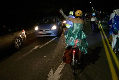A whimsical nighttime procession unfolds along Southeast Lincoln Street as costumed cyclists transform Portland's asphalt into a makeshift ballroom. Two riders in elaborate formal wear—one sporting a shimmering teal gown, the other in polka-dotted attire—navigate between curious motorists with theatrical flair. The scene captures the anarchic spirit of Pedalpalooza's Drop Out Prom Ride, where bicycle culture collides with formal rebellion under the glow of headlights and street lamps.