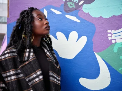 Artist Naomi Likayi stands in contemplative profile against her vibrant mural at Portland's boarded World Trade Center, her natural locs framing her upward gaze. The kaleidoscopic backdrop features flowing abstract forms in electric blues, purples, and mint greens with playful white hand motifs. Golden hoop earrings catch the light as she wears a geometric plaid coat, creating an intimate portrait that bridges the artist with her transformative public art commissioned by the Portland Street Art Alliance.