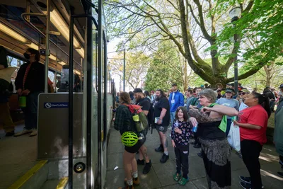 A diverse crowd of transit enthusiasts and families gathers around TriMet's retiring Type 1 MAX light rail car during its farewell event at Holladay Park in Portland's Lloyd District. The spring afternoon light filters through mature trees as passengers board for one last ride on the historic vehicle that has served the city since the system's inception. Children and adults alike press close to the train, their faces reflecting both nostalgia and curiosity as they witness the end of an era in Portland's public transportation history.