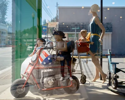 A quirky antique or thrift store window display in Vernonia, Oregon, photographed through the glass with the sunny small-town street reflected in it. The display features several vintage child mannequins posed on and around a red mini motorcycle with an American flag-patterned fairing. A female adult mannequin in a teal 1920s-style dress and white cap pushes a vintage metal shopping cart with a child mannequin seated in it. A small vintage tricycle sits to the right. The main street of Vernonia, including a green lamppost, utility poles, and storefronts, is visible in the reflection.