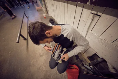 A young skateboarder crouches in concentrated focus, carefully adjusting his board's trucks in the industrial confines of Commonwealth Skateboarding in Portland's Buckman neighborhood. The warm overhead lighting casts dramatic shadows across the weathered wooden ramp structure, while concrete floors and metal railings frame this intimate moment of preparation. His gray hoodie and dark jeans blend with the utilitarian atmosphere of this Southeast Portland skate sanctuary, where craft meets passion.