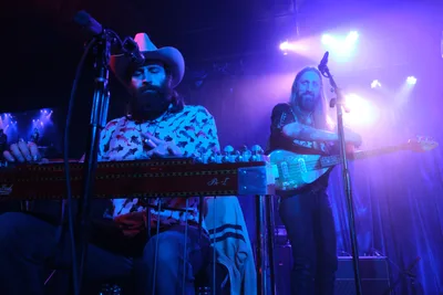 Barry Walker commands the keyboard while Tobias Berblinger anchors the rhythm on bass as Roselit Bone delivers an electrifying performance at Dante's in Portland, Oregon. The duo is bathed in saturated blue and purple stage lighting that cuts through the intimate venue's darkness, creating an atmospheric backdrop for their musical collaboration. Walker's cowboy hat and patterned shirt catch the colored light as he leans into his performance, while Berblinger's long hair flows as he works the bass strings. The dramatic lighting transforms the small club into a sonic cathedral, with beams piercing through the smoky air above the musicians.