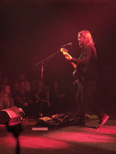 Russell Marsden of Band of Skulls emerges from crimson stage light during the band's 10 Year Anniversary Tour at Manhattan's historic Webster Hall. The guitarist-vocalist leans into his microphone with shoulder-length blonde hair catching the warm red illumination, while an array of effects pedals spreads across the venue's wooden stage floor. The intimate crowd sits in shadows beyond the performance area, creating a striking contrast between the illuminated performer and the darkened audience.