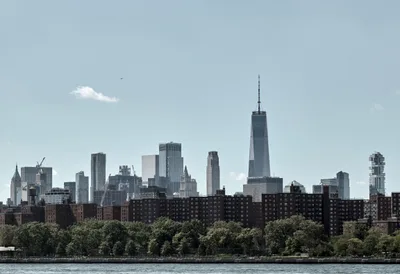 The gleaming spire of One World Trade Center pierces a hazy afternoon sky, dominating Manhattan's Financial District skyline as viewed across the Hudson River. Red-brick housing projects stretch along the waterfront in the foreground, their uniform facades creating a textural contrast against the soaring glass and steel towers beyond. Lush summer foliage softens the urban landscape while a solitary bird glides through the pale blue expanse above the city's architectural symphony.