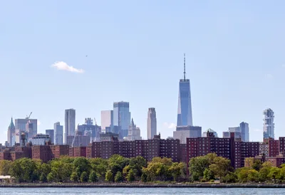 The gleaming spire of One World Trade Center pierces a hazy afternoon sky, dominating Manhattan's Financial District skyline as viewed across the Hudson River. Red-brick housing projects stretch along the waterfront in the foreground, their uniform facades creating a textural contrast against the soaring glass and steel towers beyond. Lush summer foliage softens the urban landscape while a solitary bird glides through the pale blue expanse above the city's architectural symphony.