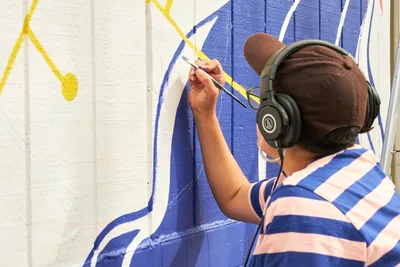 Mexican-American artist Maria Rodriguez, known as Sparkykneecap, works intently on her commissioned mural "Let's Talk" at Open Signal in Portland's Eliot neighborhood. Wearing black headphones and a blue-and-white striped shirt, she applies precise brushstrokes of vibrant blue paint to the white wall surface. The emerging artwork features bold geometric shapes and flowing yellow accents that reflect her signature exploration of identity and cultural themes through playful color and form.