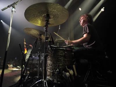 Julian Dorio commands his drum kit during Band of Skulls' 10 Year Anniversary Tour performance at the iconic Webster Hall in Manhattan's East Village. Dramatic stage lighting cuts through the venue's atmospheric haze, illuminating golden cymbals and the drummer's focused expression as he drives the rhythm forward. The intimate concert setting captures the raw energy of live rock performance, with microphone stands and equipment creating layered silhouettes against the moody backdrop.