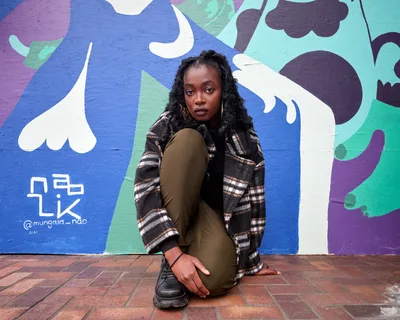Artist Naomi Likayi sits contemplatively on weathered brick pavement before her vibrant street mural at Portland's World Trade Center. Her natural curls frame her face as she gazes directly at the camera, wearing olive-green fitted clothing and a plaid flannel jacket. Behind her, abstract organic shapes in brilliant blues, purples, and greens flow across the wall, creating a dynamic backdrop that contrasts beautifully with the urban setting.
