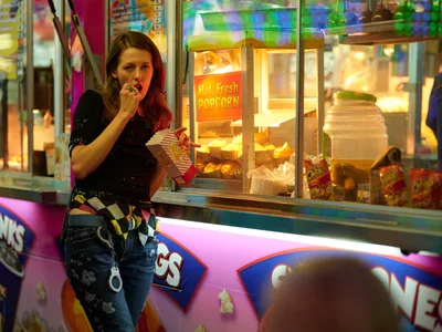 A young woman with auburn hair enjoys popcorn from a striped paper container beside a glowing concession stand at the Clark County Fair in Vancouver, Washington. The warm amber light from the "Hot, Fresh Popcorn" machine creates an inviting atmosphere against the kaleidoscope of carnival colors and neon signage. Her casual attire of distressed jeans and dark top contrasts with the vibrant fair environment, capturing a moment of simple pleasure amid the sensory overload of the midway.