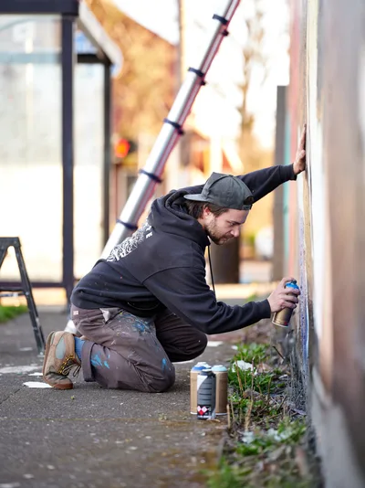 Travis Fields, known as campographic, kneels intently against a weathered concrete wall, spray can in hand as he works on his tribute mural to poet Amanda Gorman. The artist, wearing a dark hoodie and backwards cap, is captured mid-creation in Portland's Alberta Arts District, with paint-stained clothes telling the story of his artistic process. Scattered spray cans and wild grass growing from sidewalk cracks frame the intimate moment of urban art in progress.