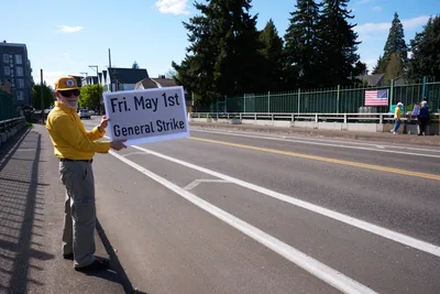 A determined activist in a bright yellow jacket and baseball cap stands on North Skidmore Street in Portland, holding a handmade sign announcing a May 1st General Strike. The bearded man positions himself against the stark urban backdrop of apartment buildings and evergreen trees under an expansive Pacific Northwest sky. His solitary but resolute stance embodies grassroots labor organizing in the quiet residential neighborhood of Humboldt.