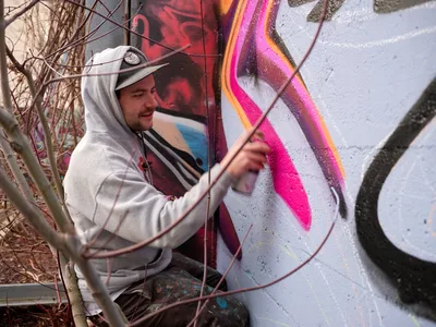 Travis Fields, known as Campographic, concentrates intently as he applies vivid magenta paint to a large-scale mural at the Clay Creative Building in southeast Portland. Framed by bare winter branches, the hooded artist works methodically on the concrete wall, his brush capturing fluid curves against a backdrop of swirling colors and abstract forms. The industrial setting and organic foreground elements create a compelling contrast between urban creativity and natural dormancy.