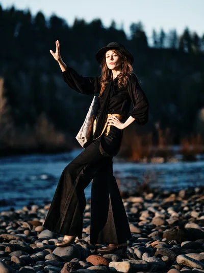 A young woman in flowing black attire and a wide-brimmed hat strikes a theatrical pose on the rocky riverbed of an Oregon waterway, one hand raised toward the sky. Warm golden hour light bathes her figure against the soft blue tones of water and distant forested hills. Her dramatic stance and bohemian styling create an artistic contrast against the natural Pacific Northwest landscape of smooth river stones and evergreen silhouettes.