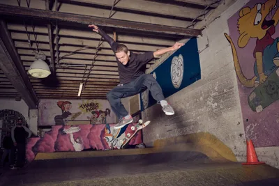 A skateboarder executes a dynamic frontside grind along a concrete ledge in the underground interior of Commonwealth Skateboarding in Portland's Buckman neighborhood. The industrial space features exposed ceiling beams, pendant lighting, and vibrant cartoon murals that create a gritty yet playful atmosphere. The skater's extended arms and focused expression capture the precision required for the technical maneuver, while colorful graffiti art transforms the raw concrete walls into a canvas of urban creativity.