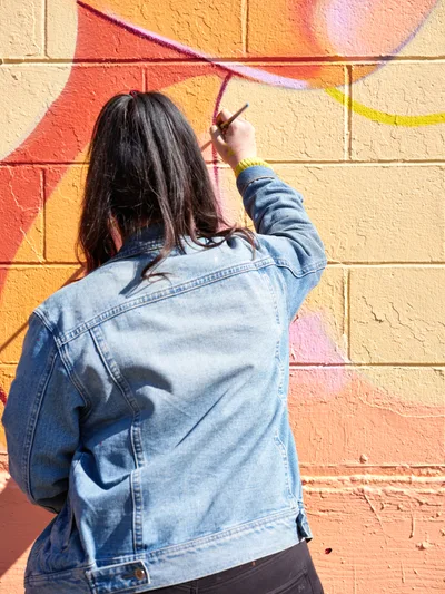 Multidisciplinary artist Vilatose works meticulously on her colorful mural 'Jardín Cubana' along Northeast Alberta Street in Portland's celebrated Alberta Arts District. Wearing a faded denim jacket with dark hair cascading down her back, she carefully applies paint to the textured brick wall, developing the three female figures that comprise the composition. The warm afternoon light illuminates both the artist and her vivid work-in-progress, capturing the creative energy that defines this historic neighborhood's artistic renaissance.