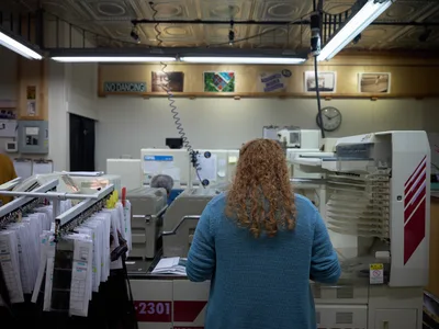 In the intimate depths of Blue Moon Camera & Machine's film processing lab on North Lombard Street, a technician with cascading curly hair works intently at specialized equipment. Fluorescent tubes cast clinical light across the cluttered workspace where vintage printing machines and drying racks create a maze of analog photography tools. The scene captures the enduring craft of film development in Portland's Saint Johns neighborhood, where traditional darkroom artistry persists amid digital dominance.
