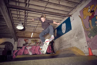 A young skateboarder launches into the air above a concrete bowl at Commonwealth Skateboarding in Portland's Buckman neighborhood, his body twisted in focused concentration. The underground space pulses with creative energy, its exposed ceiling beams and industrial pendant lights casting dramatic shadows across graffiti-covered walls featuring cartoon characters and vibrant murals. The raw concrete transitions create perfect curves for this moment of athletic poetry, captured mid-flight in a space where street culture and artistic expression converge.
