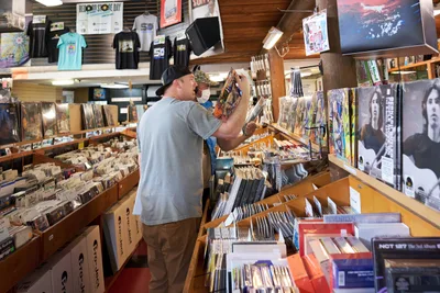 Two customers browse through extensive vinyl collections during Record Store Day 2022 at Music Millennium on East Burnside Street in Portland's Kerns neighborhood. The bearded man in a gray shirt and black cap examines album covers while his companion searches nearby bins. Warm fluorescent lighting illuminates towering wooden shelves packed with records, vintage posters, and band merchandise, creating an intimate atmosphere of musical discovery in this iconic independent record shop.