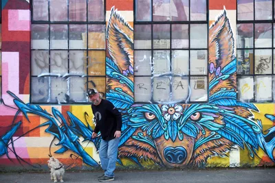 A middle-aged man in casual black attire stands confidently beside his small white terrier against a towering street art mural in Portland's Central Eastside. The vibrant artwork depicts a mystical winged wolf with piercing eyes and azure plumage, its protective gaze watching over the industrial neighborhood. Behind the mural, weathered warehouse windows reflect the soft overcast light typical of the Pacific Northwest, creating an atmospheric backdrop that bridges urban grit with artistic transformation.