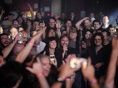 Band of Skulls members Matt Hayward and Emma Richardson perform in the middle of a packed, energetic crowd at Webster Hall in New York City in September 2019. Matt, with long hair and wearing a black shirt, holds a microphone and smiles broadly, while Emma stands beside him. Audience members surrounding them cheer, raise their hands, and hold up smartphones to capture the moment. The dimly lit venue is filled with excitement and motion blur in the foreground.