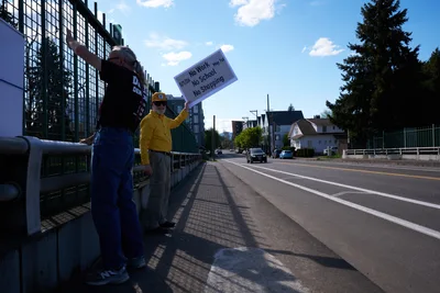 Two activists stand on North Skidmore Street in Portland's Humboldt neighborhood, holding a protest sign calling for a May 1st General Strike with the message "Stop No Work No School No Shopping." The demonstrators are positioned along Interstate 5, with one wearing a bright yellow jacket and cap while the other sports denim overalls. Late afternoon sunlight casts long shadows across the sidewalk as fluffy white clouds drift through a brilliant blue sky above the urban landscape of mixed residential and modern glass buildings.